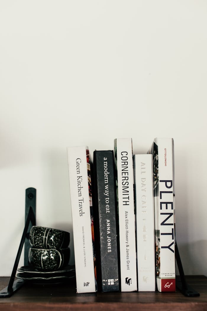 Collection of different books and cup for tea with saucers placed on wooden shelf against white wall